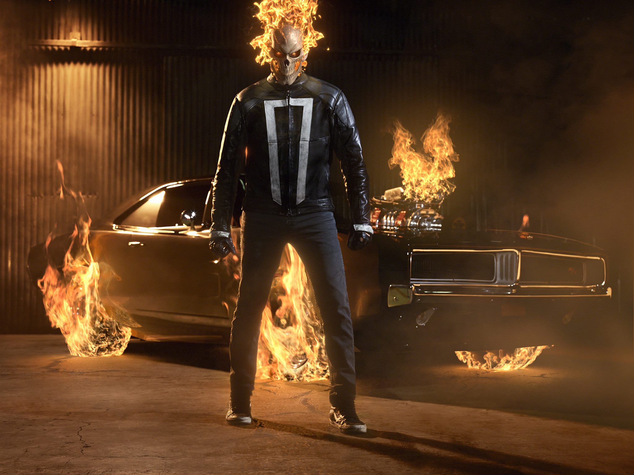 Ghost Rider with his skull on fire, standing in front of a car with the tires and engine on fire as well.