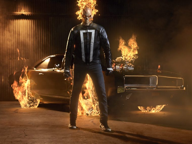 Ghost Rider with his skull on fire, standing in front of a car with the tires and engine on fire as well.